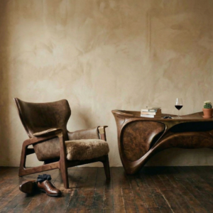 A pair of dress shoes and socks on the floor in front of a stylish wooden chair in front of a unique side table with a stack of books, car keys, a glass of wine, and a small, flowering ornamental cactus.